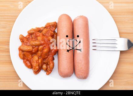 Two fat sausages with sad faces, lying on a white plate with a fork and beans, conceptual image about diet, healthy eating and obesity Stock Photo