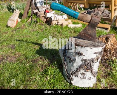 A wooden deck from a birch trunk with a stuck ax with a rewound blue electrical tape handle against a background of chopped firewood and green grass. Stock Photo