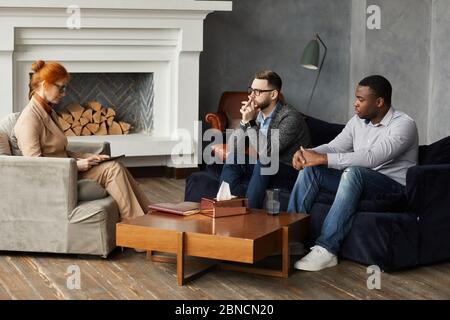 Two serious men sitting on sofa and listening to conclusion of psychologist during therapy at office Stock Photo