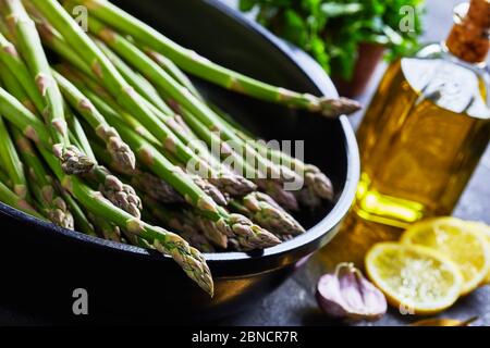 close-up of fresh green asparagus in black baking dish with garlic, lemon slices, peppercorns and bottle of olive oil on the grey concrete table, hori Stock Photo