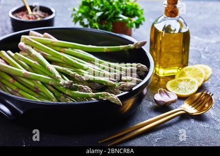 close-up of fresh green asparagus in black baking dish with golden cutlery, lemon slices, peppercorns and bottle of olive oil on the grey concrete tab Stock Photo