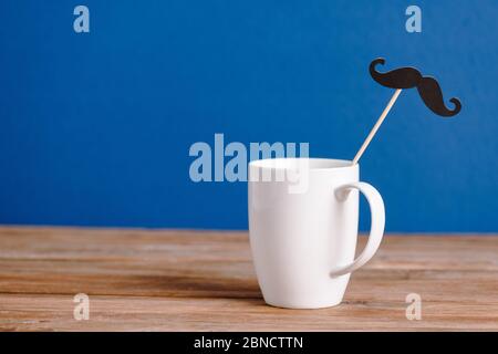 white cup and black decorative paper fake mustache on wooden surface isolated on blue Stock Photo