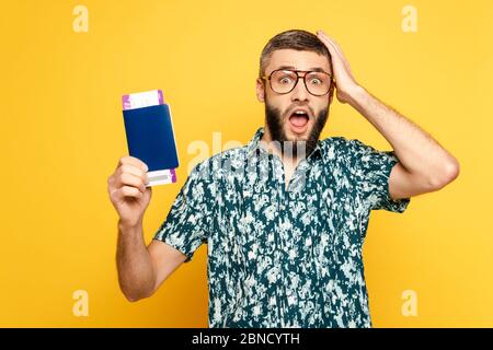 shocked bearded guy in glasses with passport and ticket holding head on yellow Stock Photo