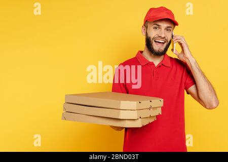 smiling bearded delivery man in red uniform with pizza boxes talking on smartphone on yellow Stock Photo