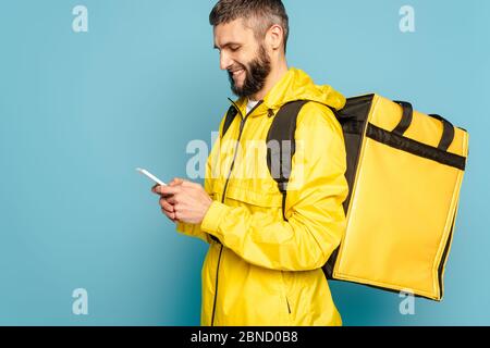 smiling deliveryman in yellow uniform with backpack using smartphone on blue background Stock Photo