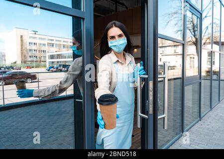 Cafe owner in medical mask near door showing disposable cup of coffee and looking at camera on street Stock Photo