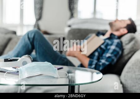 Selective focus of medical mask and headphones on coffee table near man with book sleeping on couch Stock Photo