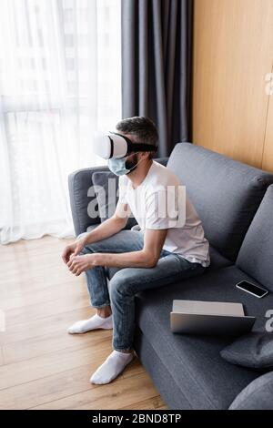 Side view of man in medical mask using virtual reality headset near gadgets on sofa Stock Photo