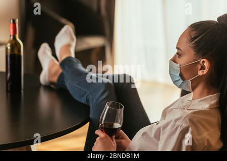 selective focus of depressed young woman in medical mask holding glass of red wine while sitting with legs on table