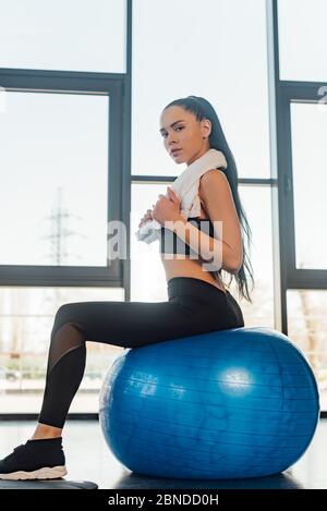 Side view of sportswoman with towel sitting on fitness ball and looking at camera in gym Stock Photo