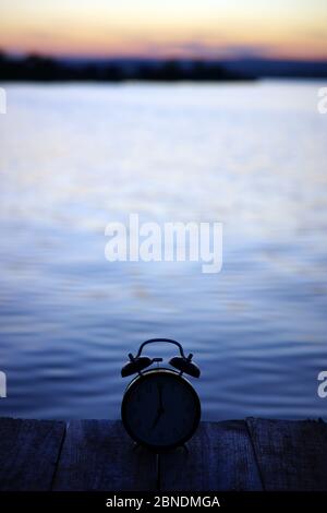 Summer time concept. Alarm clock silhouette on wooden bridge on sea sunrise background Stock Photo