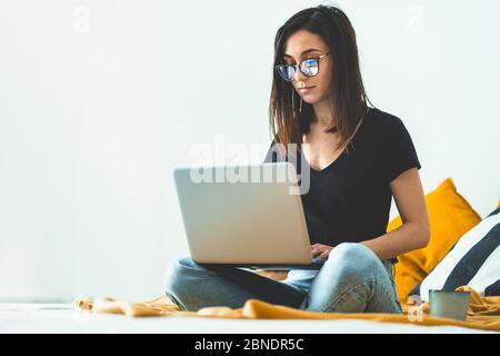 Attractive young woman wearing eye glasses working from home. Female entrepreneur sitting on bed and working from laptop computer while drinking morni Stock Photo