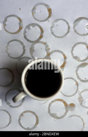 A white cup of black coffee sits on a white paper tablecloth background covered in rings of wet coffee stains Stock Photo