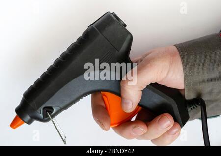 Hand holds an electric glue gun. Man in gray protective clothing. Using a hand tool. Close-up. Selective focus. Stock Photo
