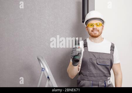 Male carpenter builder is holding electric screwdriver in hands on stepladder in safety glasses and helmet Stock Photo