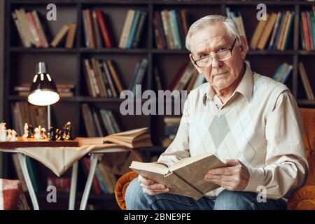 Senior man alone sitting at library in glasses holding book thoughtful Stock Photo