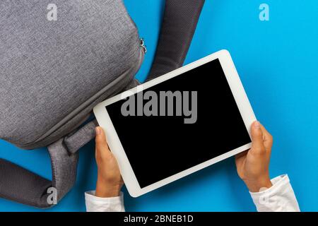 Kid hands with digital tablet computer and gray backpack over light blue background. Top view Stock Photo