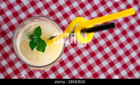 A glass of lemonade with green mint leaves and two straws on a checkered red-and-white tablecloth. Stock Photo