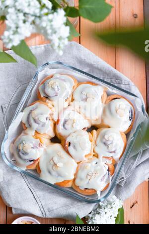 Top view of fresh tasty cinnamon rolls buns with cream and almonds on a wooden table with white turns of lilac on a sunny day, vertical photo Stock Photo