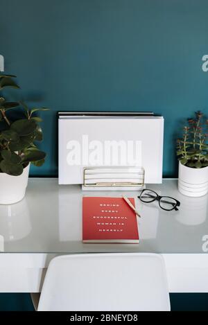 Closeup view of a white desk with white papers, red notebook, plants, pen and glasses in it Stock Photo