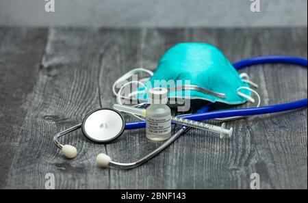 A blue medical stethoscope, face mask respirator, syringe and vial of covid-19 test vaccine on a wooden surface. Stock Photo