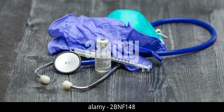 A blue medical stethoscope, face mask respirator, gloves, syringe and vial of covid-19 test vaccine on a wooden surface. Stock Photo