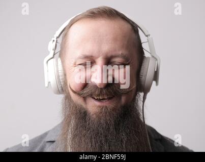 Man with a long beard and a mustache wearing white earphones, standing in front of a grey wall Stock Photo