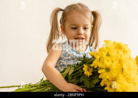 childhood, holidays, flowers, gifts concept - little cute three year old girl with two ponytails on her head in blue colorful dress holds large Stock Photo