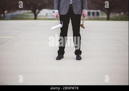 Man in a suit holding a paint roller and hammer standing on paved ground Stock Photo