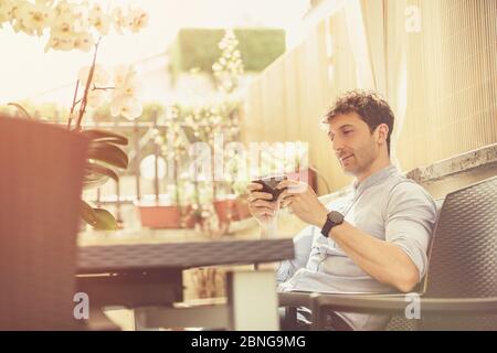 Outdoor man sitting and relaxed texting with a smartphone in his hands. Illuminated environment, man wears a heavenly shirt. Spring scene. Social conn Stock Photo