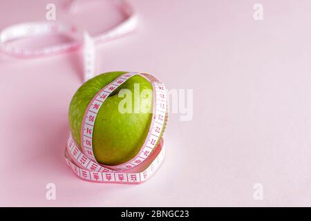 selective focus, green ripe Apple wrapped in measuring tape, the concept of proper nutrition and weight loss. on a pink background Stock Photo