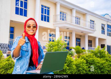 pretty young black lady sitting alone with her laptop computer outdoor in a park, giving thumbs up and smiling Stock Photo