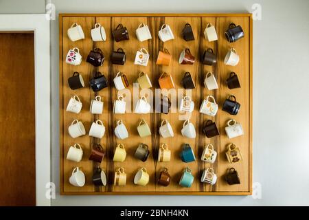 View of a variety of colorful and beautiful mugs hanging from a wooden surface on a white wall Stock Photo