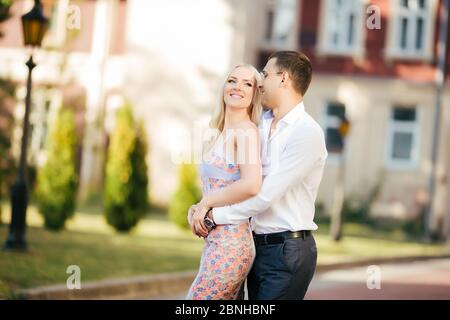 Lovely couple walking around the block. Dark-haired man in a white shirt hugging a blonde in a beautiful dress Stock Photo