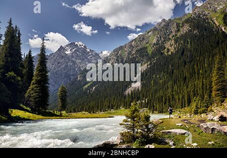 Tourist with big backpack is walking down the road in the forest of mountain valley in Karakol national park, Kyrgyzstan Stock Photo