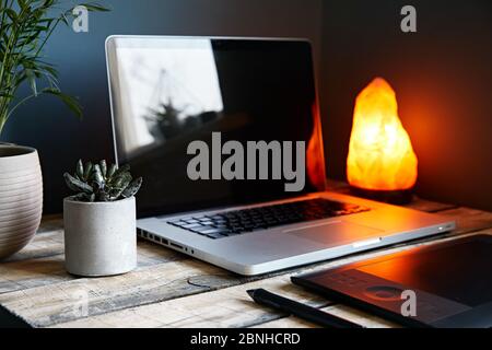 Cozy workspace with laptop, graphic tablet, plants and yellow glowing lamp at stone grey wall background. Working from home concept. Stock Photo