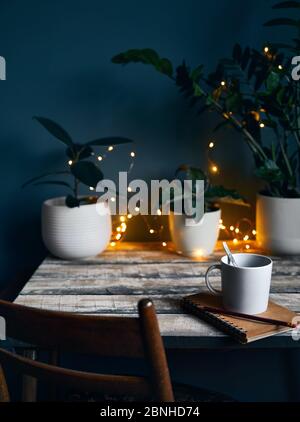 Cozy workspace with note book with and cup of coffee at wooden table with plants and yellow glowing garland at stone grey wall background. Working fro Stock Photo