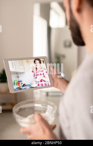Over shoulder view of mature mother with bowl of flour teaching adult son to cook while talking to him via telecommunication app in isolation Stock Photo