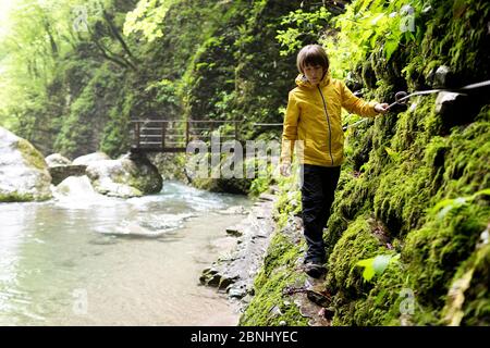 Young boy in yellow jacket walking through river gorge holding a safety rope to Kozjak waterfall, Slovenia Stock Photo