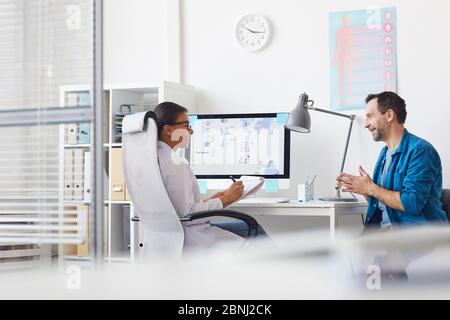 Young smiling doctor sitting at her workplace talking to her patient and making notes in medical card at office Stock Photo