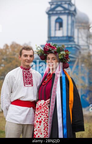 Girl and guy in retro costumes on the street in the old village. Retro staging of an ancient rite. Beautiful wreath on a girl. The guy hugs the girl Stock Photo