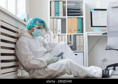 Doctor in protective clothing sitting on the floor at office and resting after hard work he using mobile phone Stock Photo