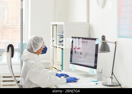 Female doctor in protective clothing sitting at the table in front of the computer monitor and examining the statistics about the coronavirus Stock Photo