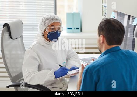 Female doctor in protective clothing making notes in document while listening to the patient Stock Photo