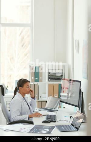 Young female doctor in white coat sitting at her workplace and working on computer at the office Stock Photo