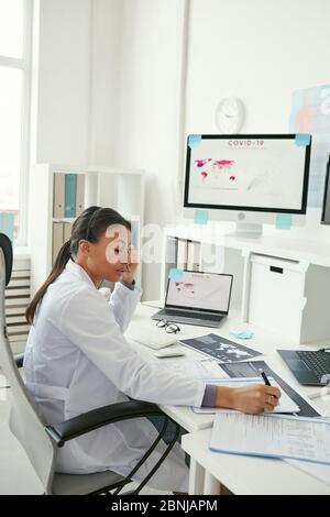Young female doctor sitting at the table doing paperwork and working on computer at her office Stock Photo