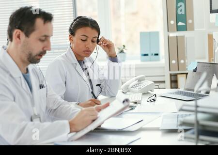 Mature male doctor in white coat making notes in document while nurse sitting at the table and taking a call at hospital Stock Photo