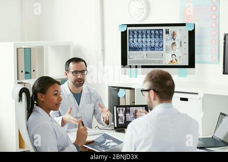 Team of doctors sitting at the table and discussing x-ray images together during online conference with their colleagues Stock Photo