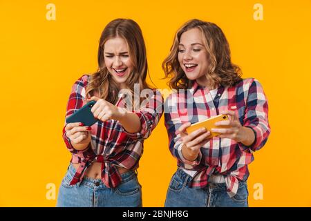 Image of two young beautiful girls wearing plaid shirts having fun and playing video games on cellphones isolated over yellow background Stock Photo