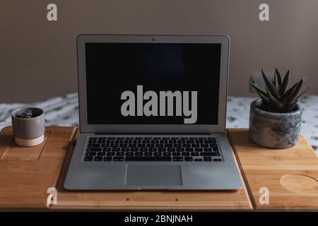 Front view of a laptop with a blank screen with house plants either side on a portable folding wooden desk on top of a bed with white patterned sheets Stock Photo